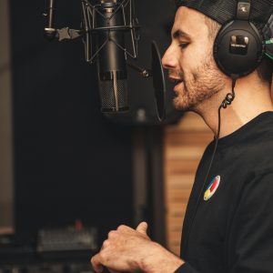 A white male stood up with a hat on speaks into a black microphone and has headphones on