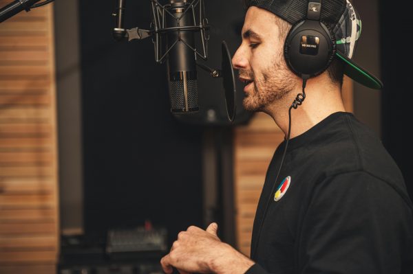 A white male stood up with a hat on speaks into a black microphone and has headphones on