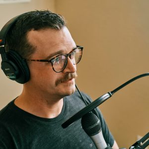 A male with glasses and a moustache speaking into a microphone in a studio