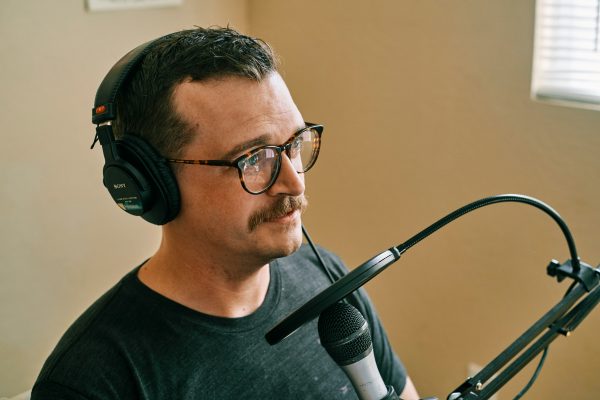 A male with glasses and a moustache speaking into a microphone in a studio